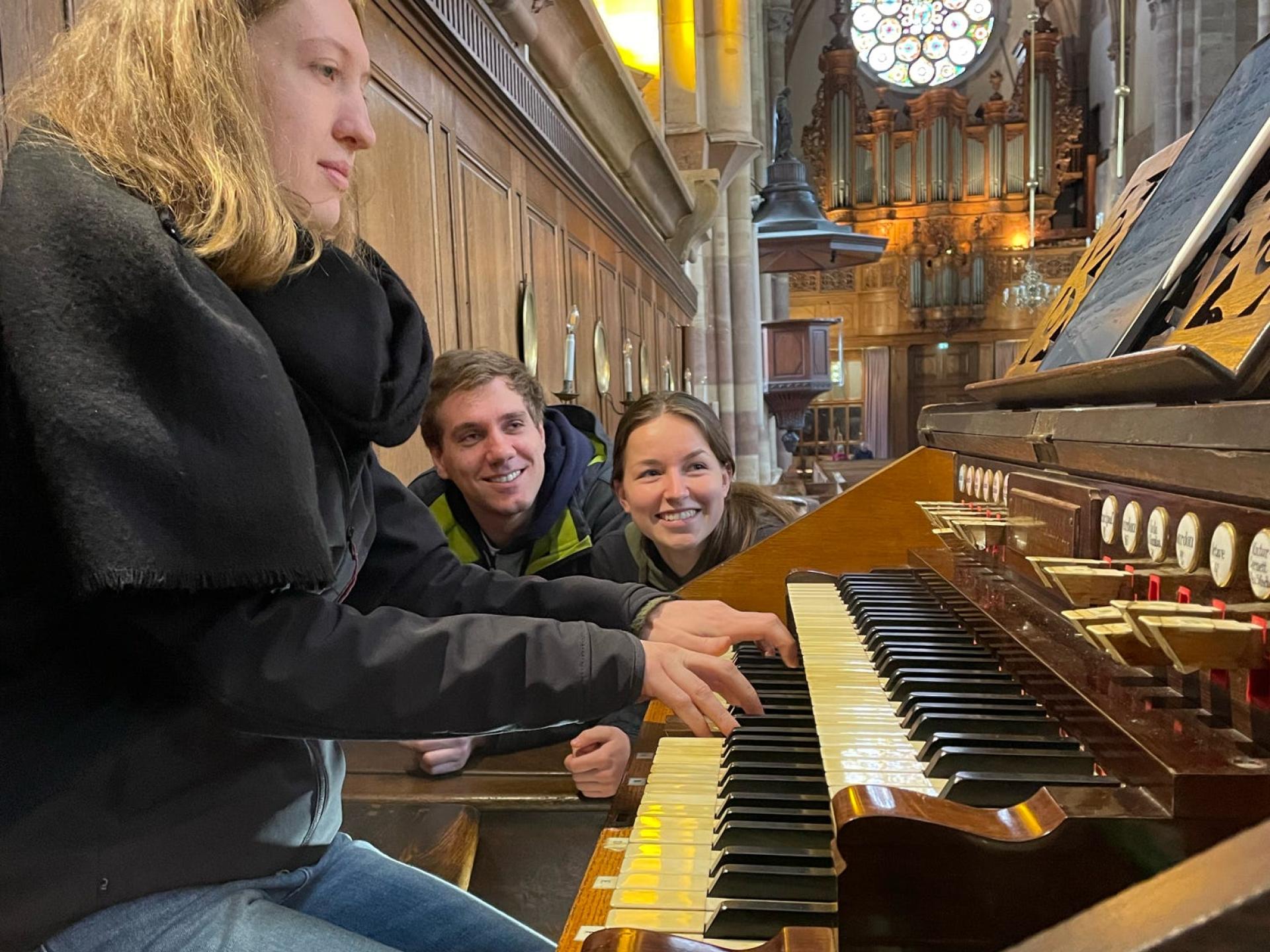 eine Studentin spielt an einer Orgel, zwei Studierende schauen ihr zu