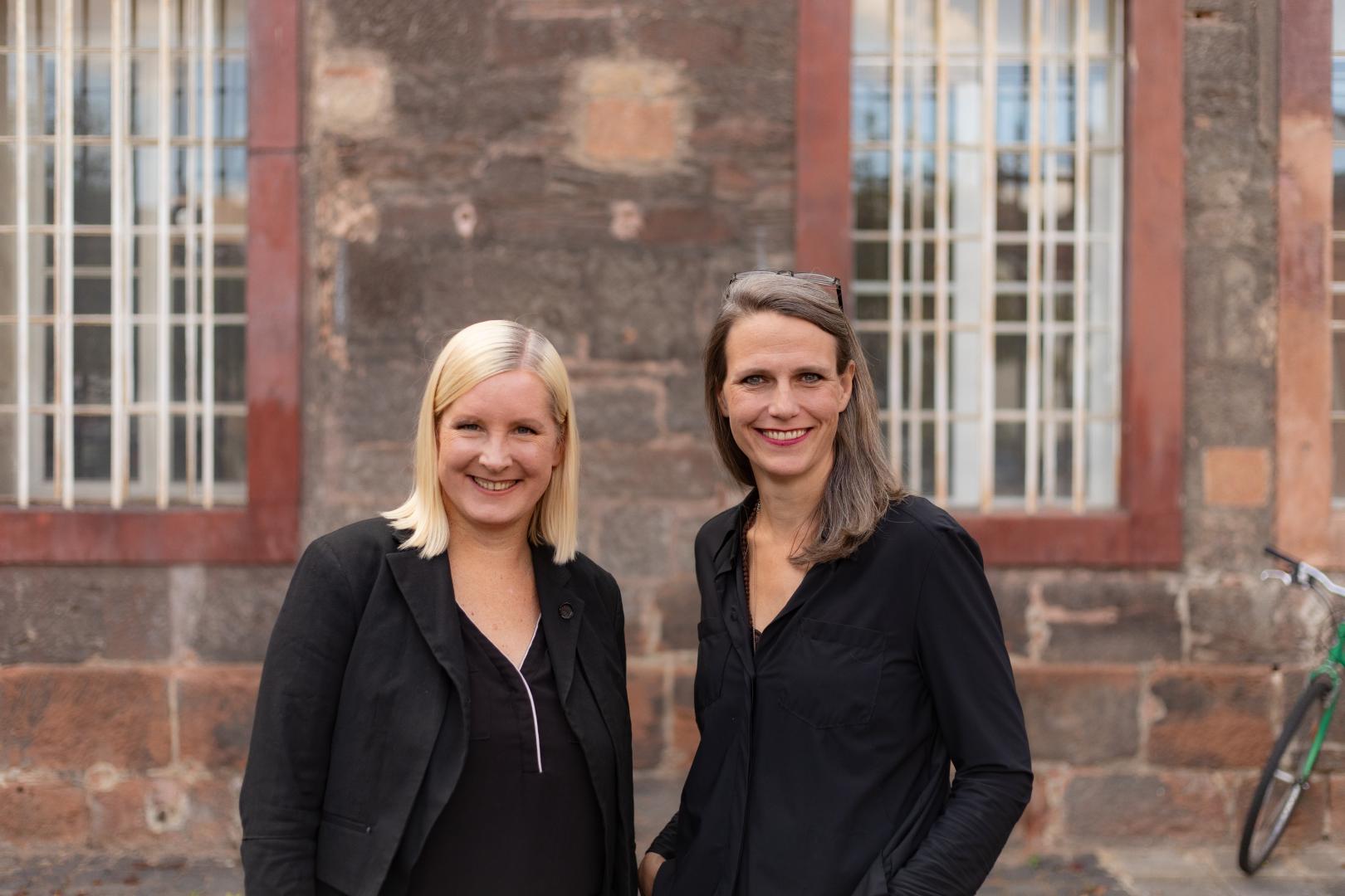 Eva Lange und Carola Unser stehen vor der rötlichen Steinwand eines Gebäudes.