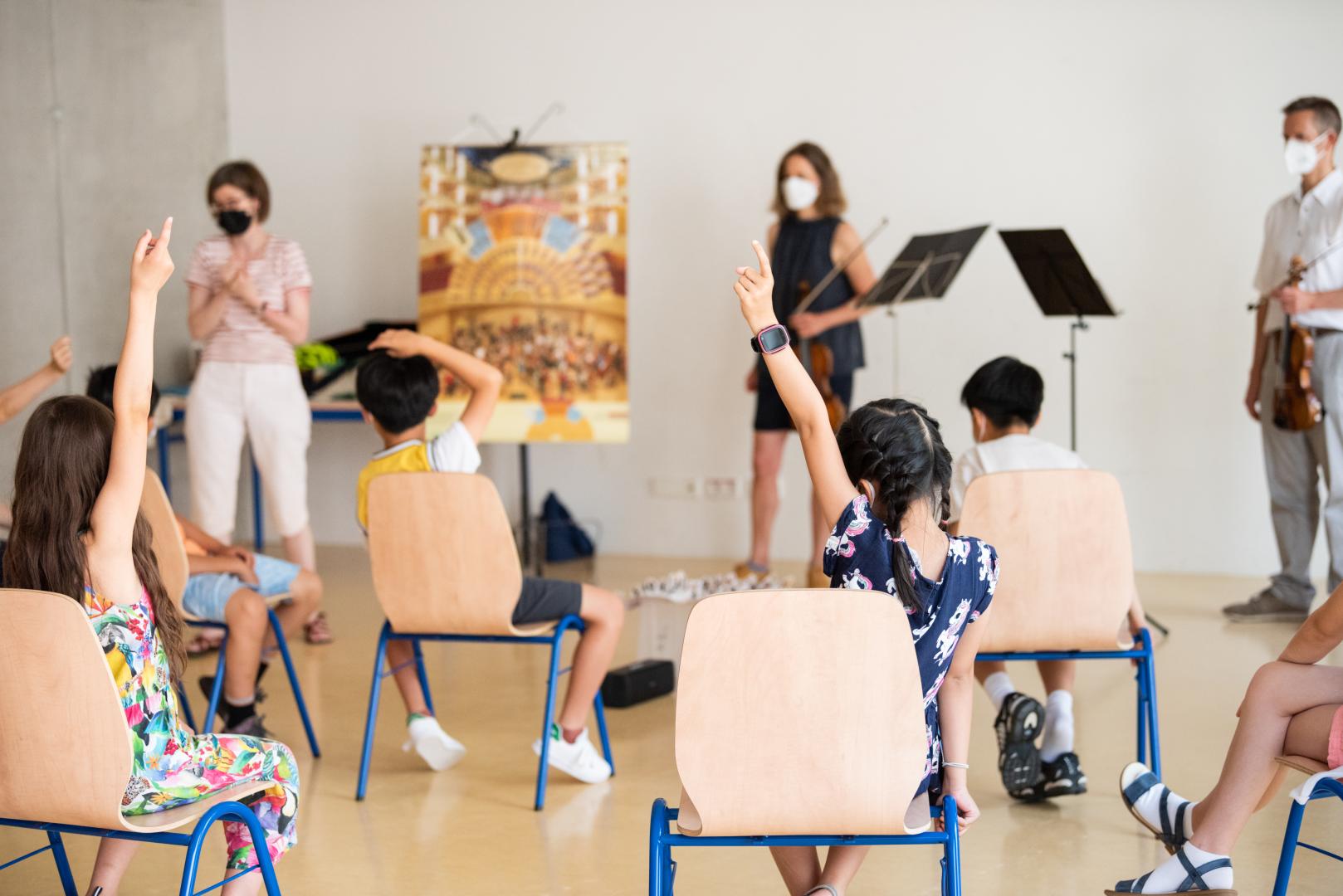 Musikprojekt mit einer Schulklasse, zwei Schülerinnen melden sich, sie sind im Vordergrund von hinten zu sehen.