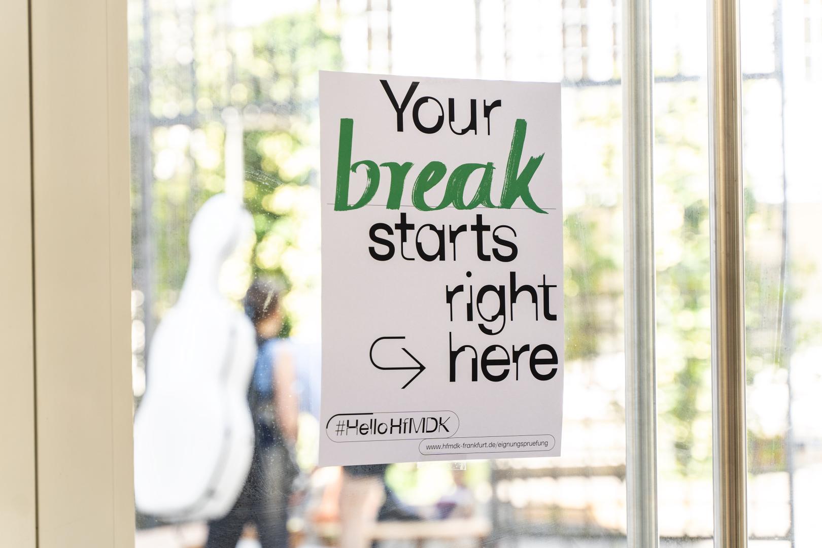 Plakat mit der Aufschrift "Your break starts right here" an der Glastür zum Innenhof der HfMDK