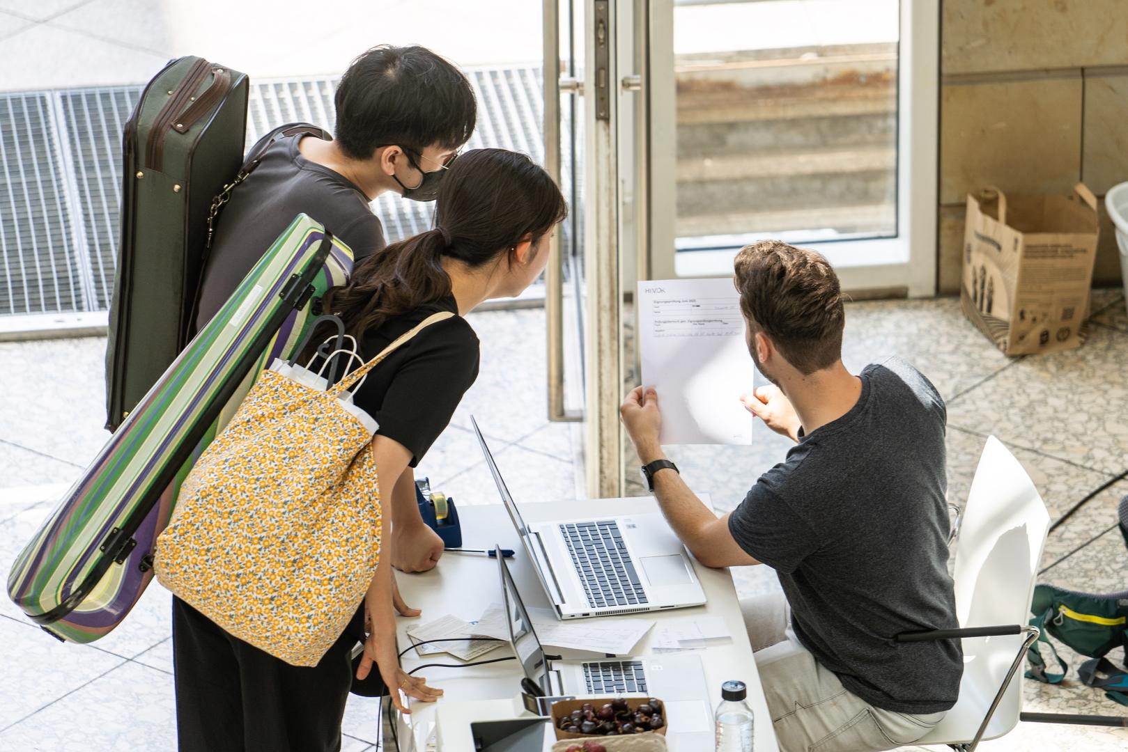 Zwei Studienbewerber*innen werden im Foyer der HfMDK von einem studentischen Mitarbeiter zur Eignungsprüfung beraten.