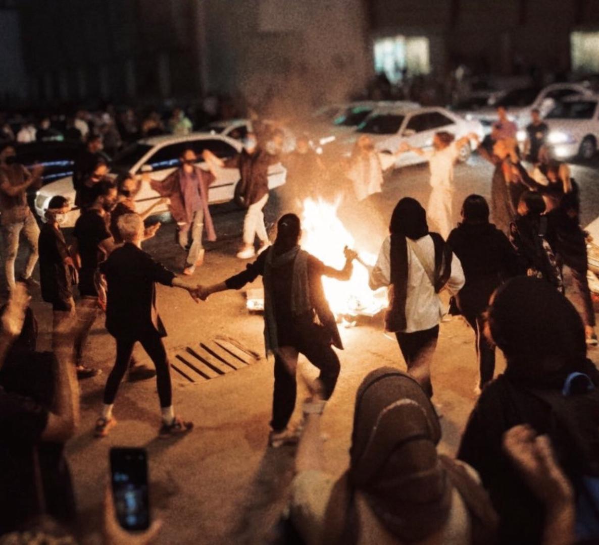 Frauen tanzen auf einer Straße um ein Feuer herum, im Hintergrund Autos.