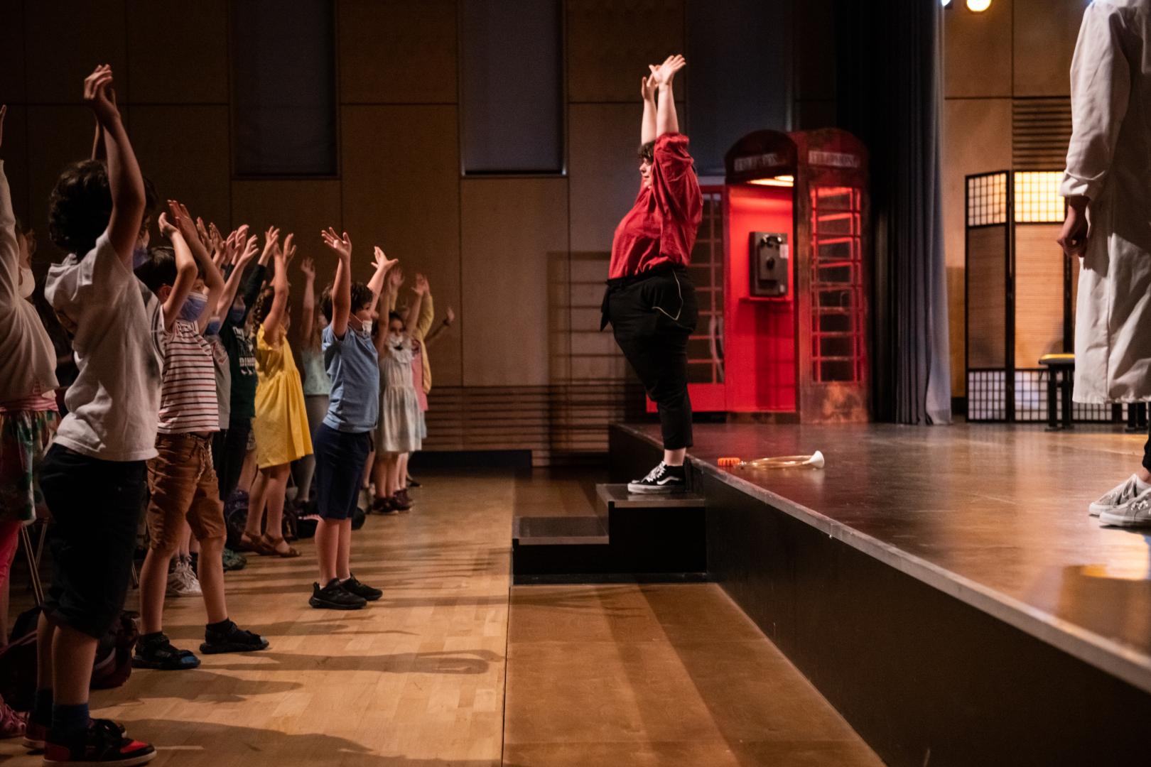 Mitmachkonzert für Kinder. Rechts auf der Bühne macht eine Musikerin vor, wie die Kinder die Arme heben sollen. Links im Bild Kindergruppe, die mitmacht.