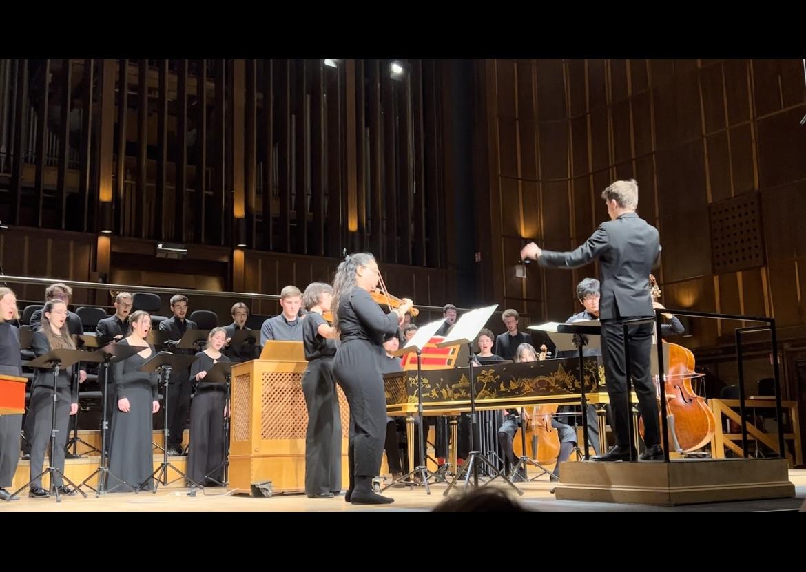 Blick von Seite auf Bühne; Student dirigiert Chor und Instrumentalist*innen