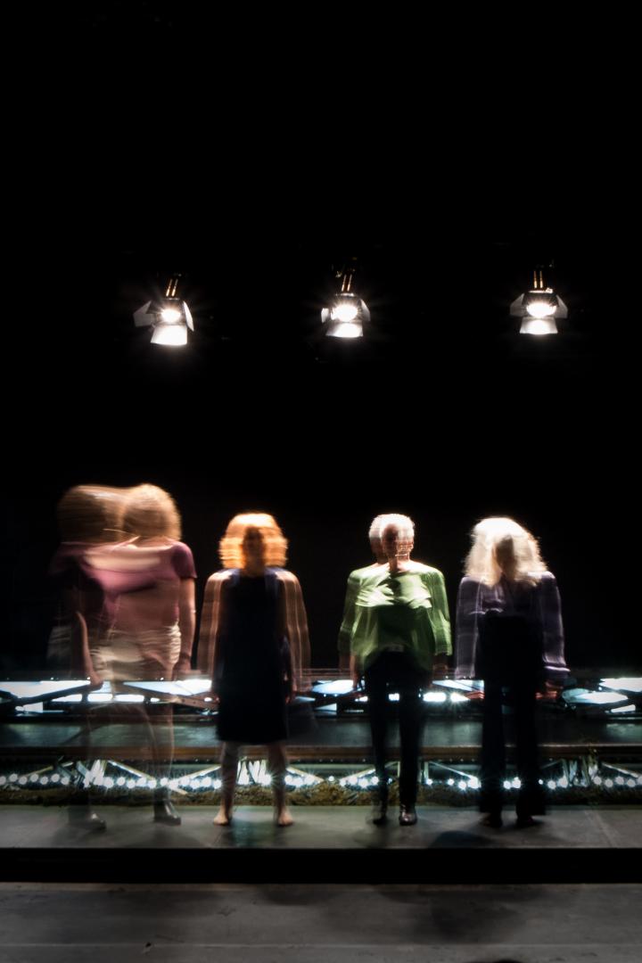 Vier Frauengestalten sind im Ganzkörper in Bewegungsunschärfe auf einer Bühne stehend zu sehen, ihre Gesichter sind nicht zu erkennen.