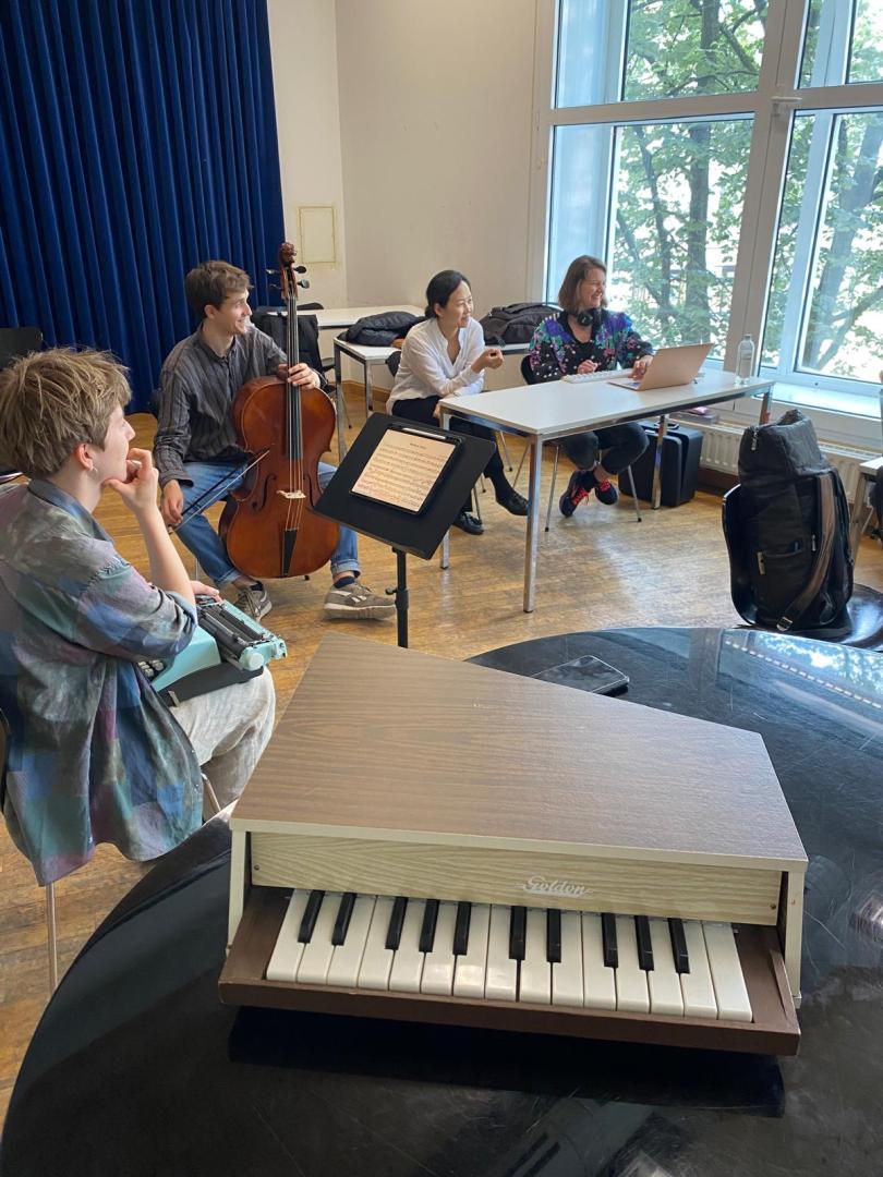 Vier Studierende sitzen in einer Probe in einem Halbkreis mit einer Schreibmaschine, einem Cello und einem Laptop. Sie lachen. Im Vordergrund setzt ein Mini-Klavier auf einem Flügel.