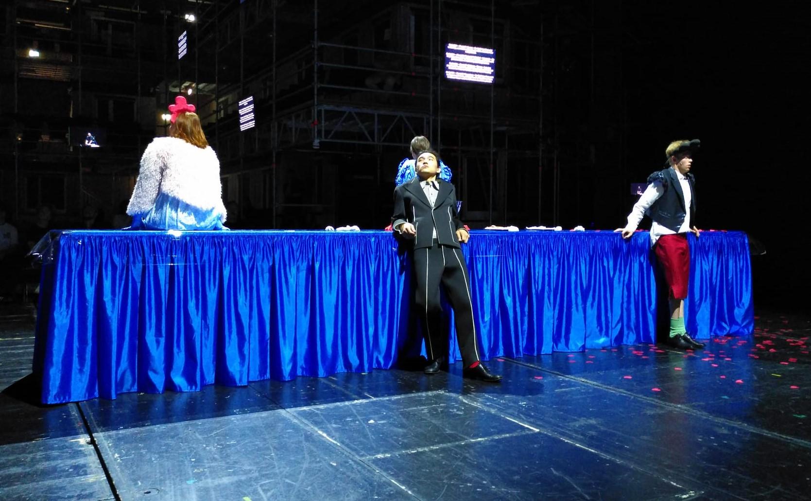 Auf einer Bühne steht ein ca. fünf Meter langer und ein Meter hoher schmaler Tisch, der von einer bodenlangen blauen Satindecke bedeckt ist. Links darauf sitzt eine Person, die von hinten zu sehen ist, mit schulterlangem rötlichem Haar und einer weißen Felljacke. In der Mitte vor dem Tisch steht ein Mann im schwarzen Anzug, er schaut gelangweilt nach rechts links oben. Direkt dahinter sitzt eine weitere Person auf dem Tisch mit dem Rücken zur Kamera.  Weiter rechts vor dem Tisch steht ein zweiter Mann.