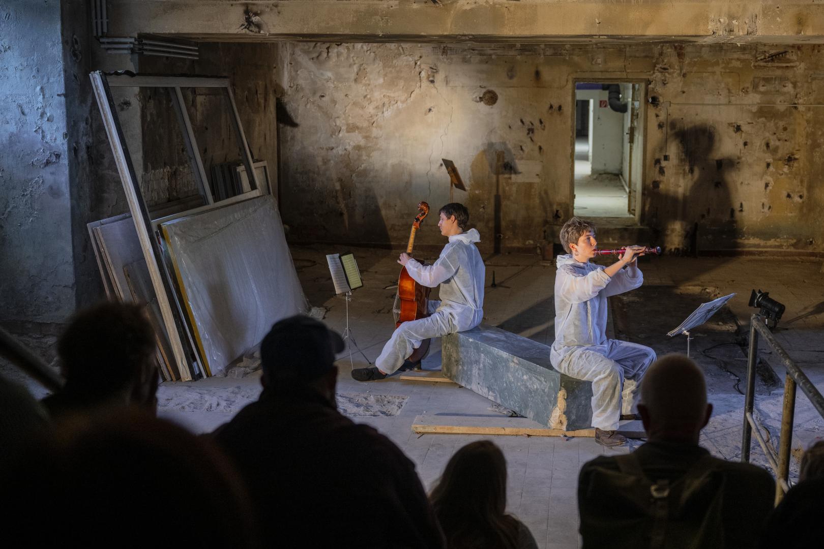 Ein Cellist und eine Blockflötistin sitzen auf einer Art rechteckigem Stein in einem baufällig aussehenden Keller und spielen. Im Anschnitt sind Zuschauer*innen zu sehen.