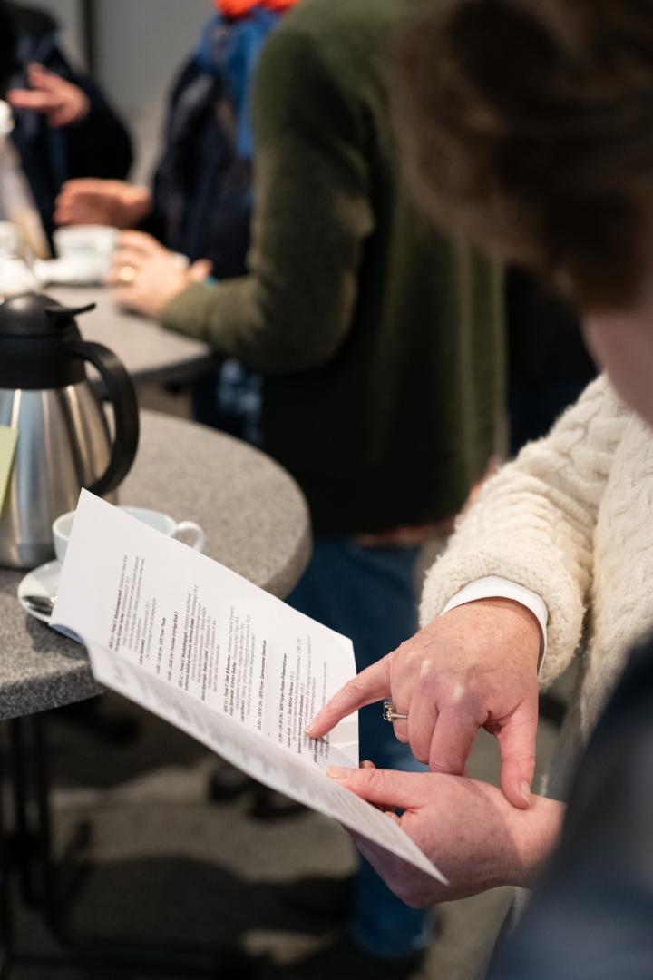 Zwei Personen betrachten einen aufgeschlagenen Programmzettel, sichtbar sind dabei nur Hände: Die linke Hand der ersten Person hält das Blatt, die andere Person deutet mit dem kleinen Finger der rechten Hand auf einen Programmpunkt. Im Hintergrund sind andere Personen und Stehtische erkennbar, auf einem der Tische steht eine Kaffeekanne.