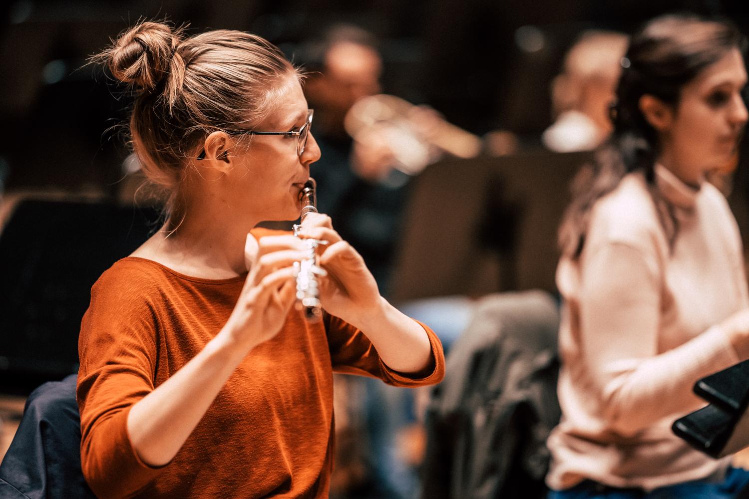 Sina Merkel spielt Querflöte in einer Orchesterprobe. Sie trägt ein organgenes Shirt, eine Brille und hat ihre dunkelblonden Haare zu einem Dutt gebunden. Man sieht sie hier im Profil.