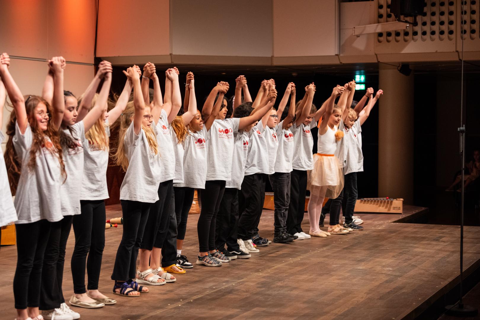 Eine Reihe von Kindern in weißen T-Shirts steht auf der Bühne und verbeugt sich. Alle fassen sich an den Händen und haben die Arme nach oben gestreckt.