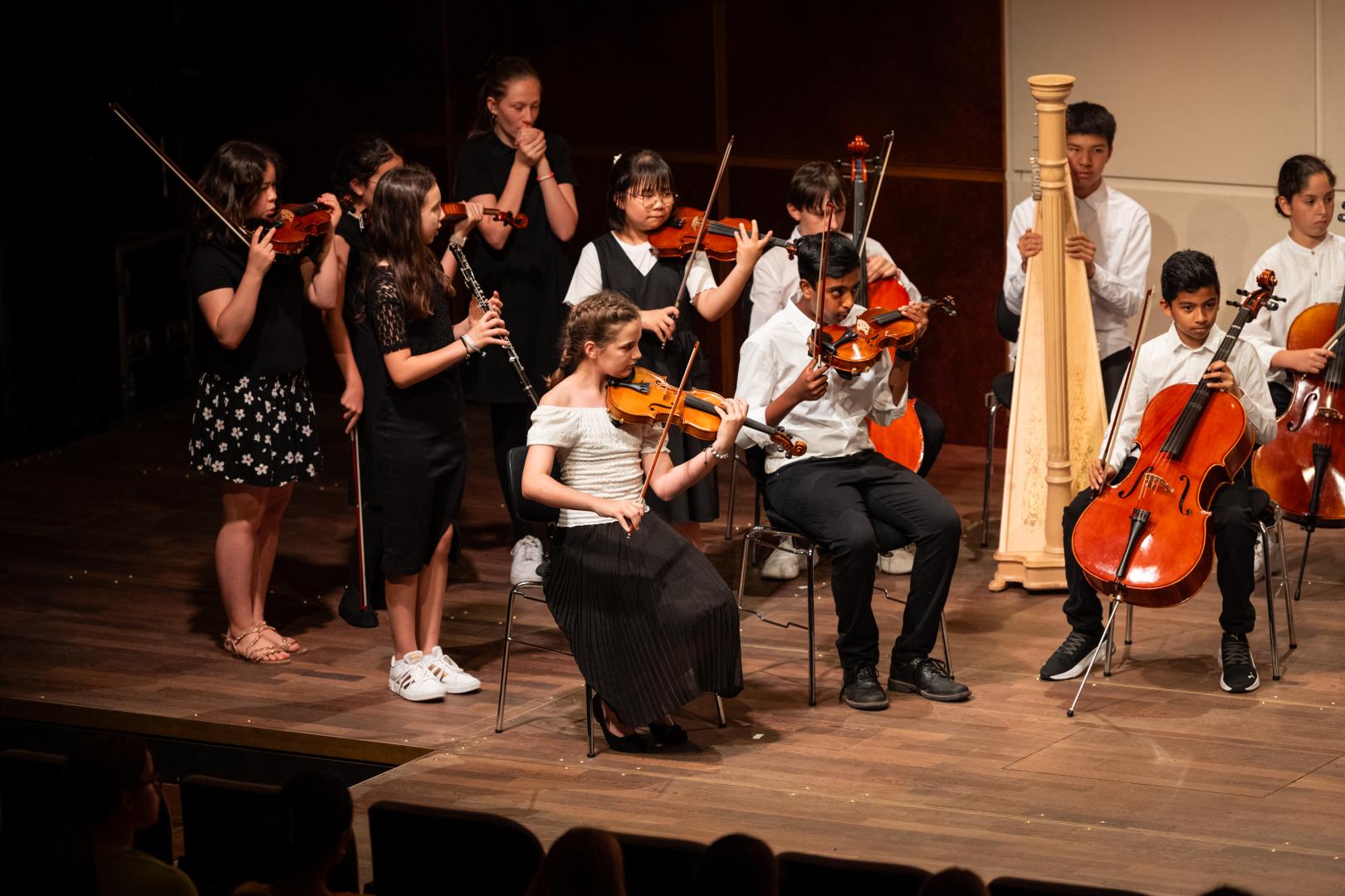 Eine Gruppe Kinder in Orchesterformation auf der Bühne. Die meisten haben Streichinstrumente. Sie tragen feine schwarz-weiße Kleidung und haben konzentrierte Gesichter. Manche von ihnen spielen gerade, die anderen scheinen auf ihren Einsatz zu warten.