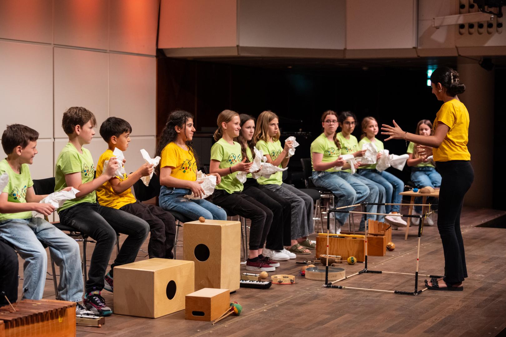 Kinder in hellgrünen und gelben T-Shirts sitzen in einer Reihe auf Stühlen und halten zerknülltes Papier in der Hand. Vor ihnen steht eine Dirigentin in einem gelben Shirt. Auf der Bühne liegen außerdem verschiedene Percussion-Instrumente.