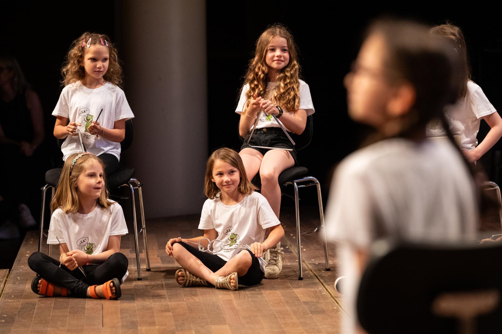 Eine Gruppe Kinder in weißen T-Shirts auf der Bühne. Zu sehen sind hier vier Mädchen. Zwei sitzen auf Stühlen, die anderen beiden davor auf dem Boden. Drei von ihnen halten je eine Triangel. Das vierte Mädchen ein ähnliches Instrument mit drei ineinandergreifenden Ringen. Alle vier blicken lächelnd nach rechts.