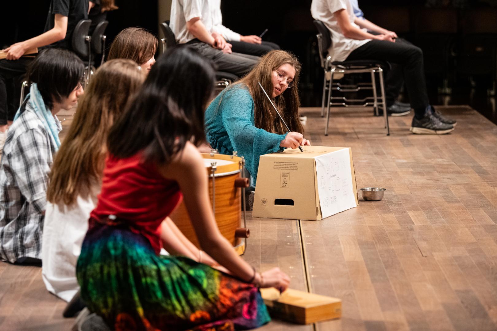 Kinder knien auf dem Boden und spielen auf mehr oder weniger improvisierten Percussion-Instrument. Im Fokus steht ein Mädchen, das mit einem Stock über einen Pappkarton streicht.