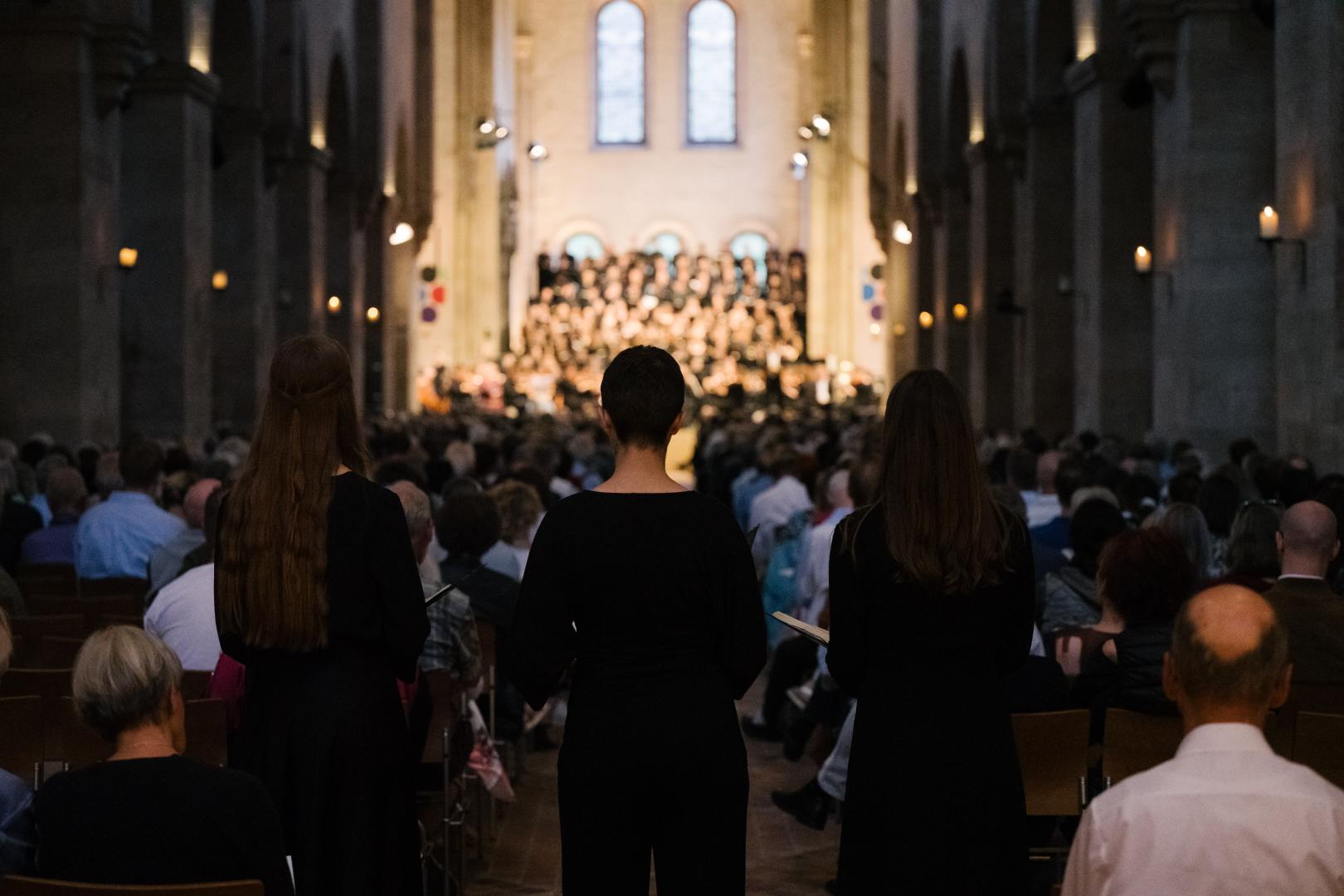 3 Studentinnen von hinten fotografiert, im Hintergrund sieht man verschwommen Chor und Orchester auf der Bühne im Kloster Eberbach