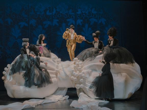 Sechs Darsteller*innen in schwarzen barocken Kostümen und eine Darsteller*in in einem goldenen Kostüm mit Trompete stehen auf einer weißen großen Wolke auf der Bühne.