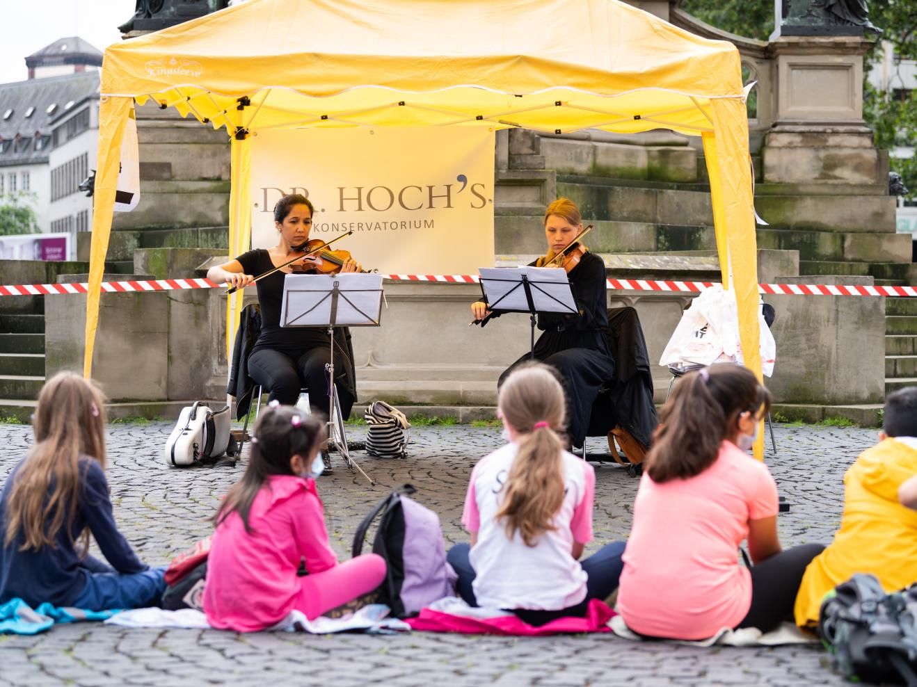 Zwei Musikerinnnen spielen unter einem gelben Pavillon, davor auf dem Boden sitzen mehrere Kinder.