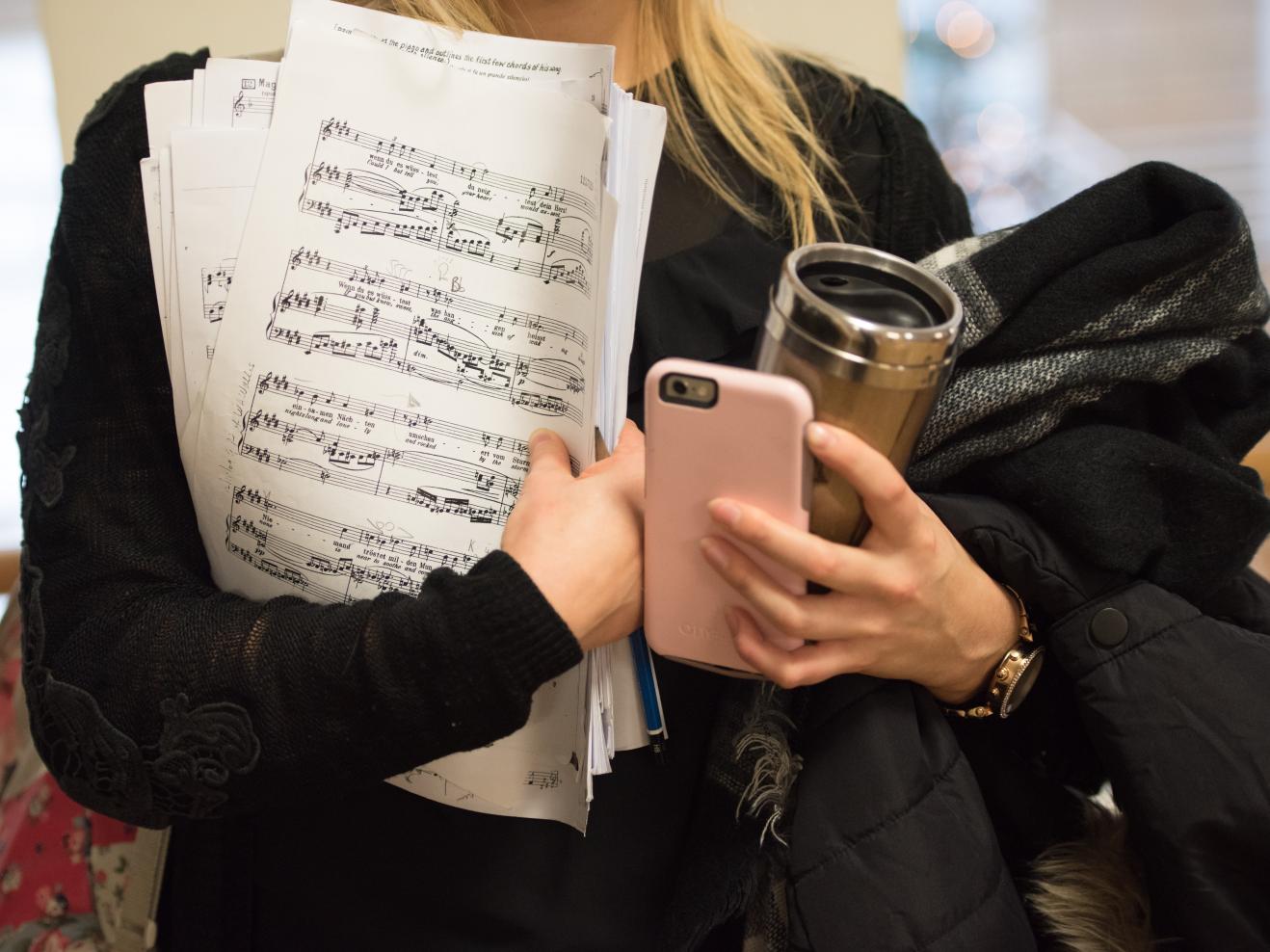 Eine Studentin hält Noten, einen Kaffeebecher, eine Jacke und ihr Smartphone in der Hand.