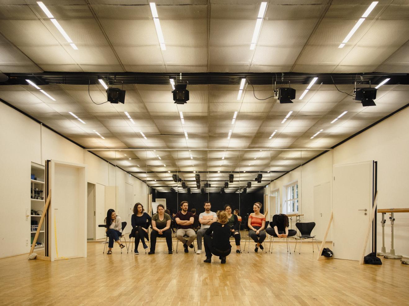 Acht Gesangsstudierende sitzen nebeneinander in einer Stuhlreihe im Opernstudio vor ihnen hockt eine Person