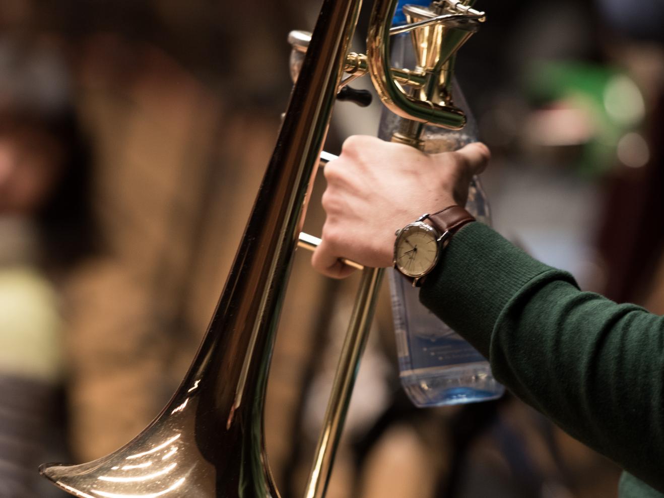 Nahaufnahme einer Posaune und einer Wasserflasche in der Hand eines Musikers, er trägt eine Armbanduhr.