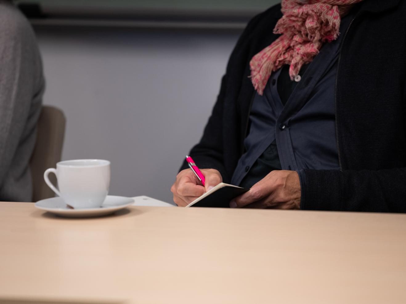 Person notiert etwas in ein Büchlein, daneben eine Kaffeetasse