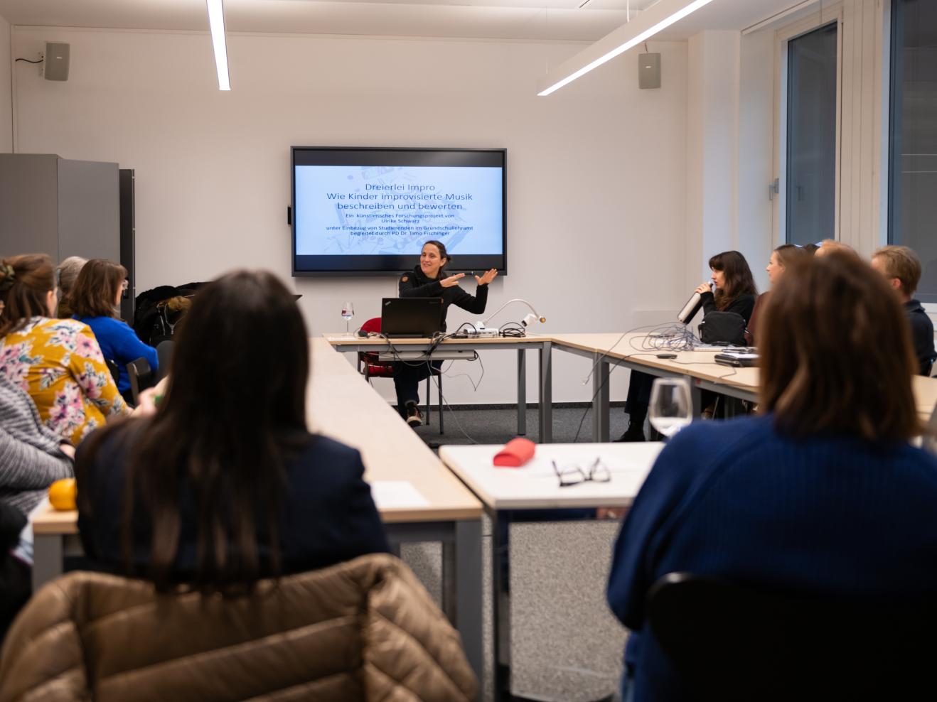 Im Vorder- und im Mittelgrund ist eine Gruppe von mehreren Zuhörenden sitzt an Tischen im Kreis. Fokussiert im Zentrum des Bildes ist eine Vortragende, die im Hintergrund an einem Tisch sitzt und mit den Händen eine erklärende Geste ausführt. Hinter der eine Präsentation auf einem Bildschirm erkennbar wird: Dort ist zu lesen: „Dreierlei Impro – Wie Kinder improvisierte Musik beschreiben und bewerten“.