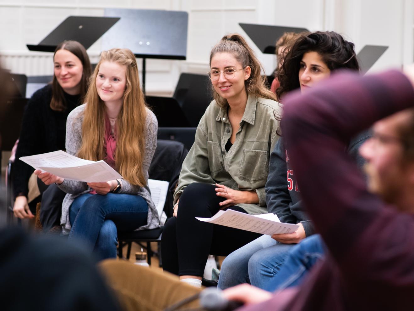 Studierende sitzen in einem Stuhlkreis. Sie hören aufmerksam zu und lächeln dabei. Die meisten haben Noten in der Hand. Eine Studentin lächelt in die Kamera.