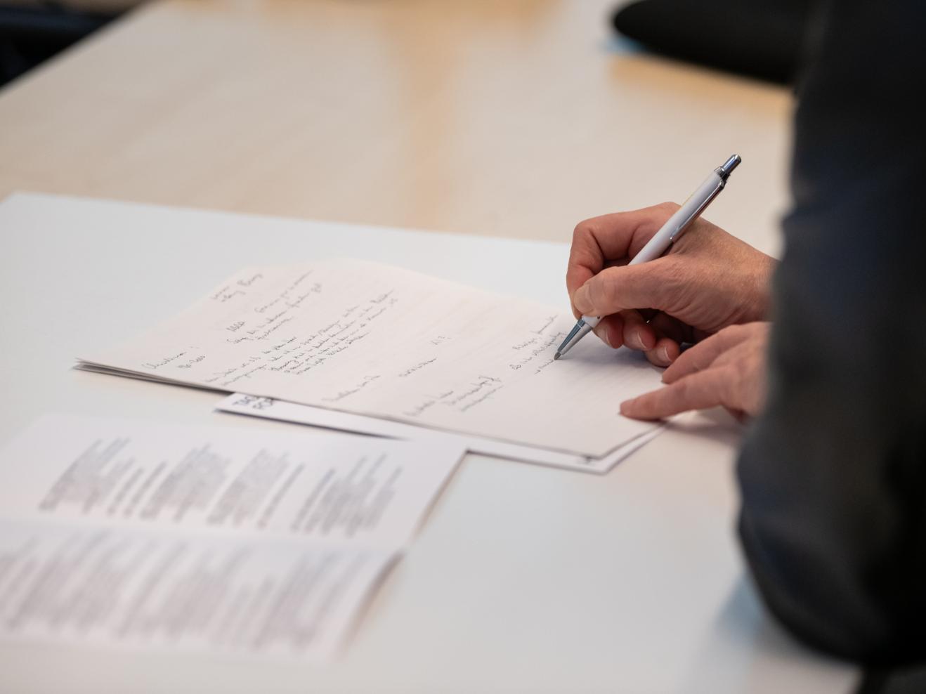 Eine Person macht sich handschriftliche Notizen auf einem Zettel auf einem Tisch, auf dem auch ein Tagungsprogramm aufgeschlagen liegt. Von der Person sind durch den gewählten Bildausschnitt nur die Hände und ein Arm sichtbar.