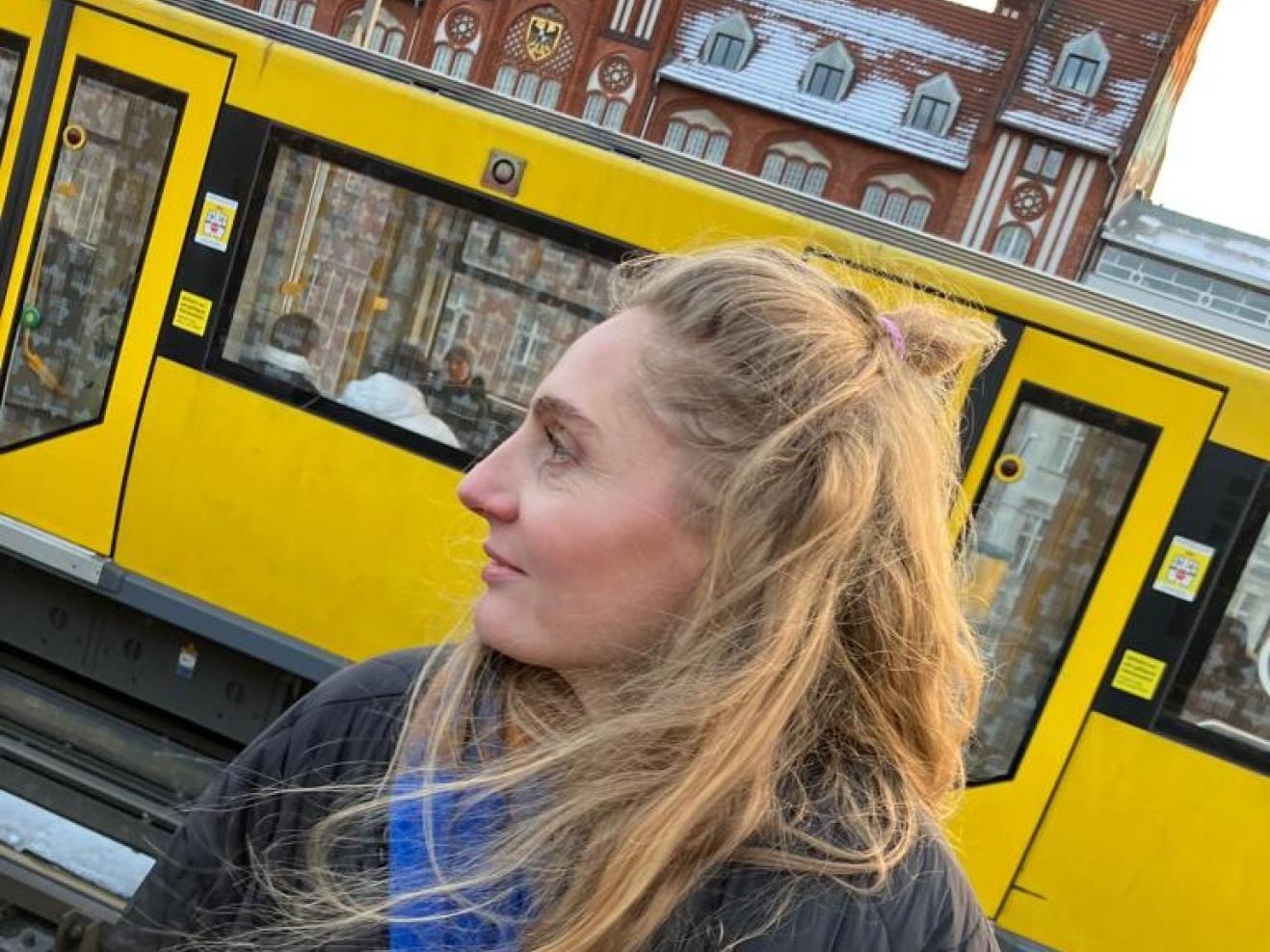 Eine Frau steht im Vordergrund, sie ist bis zur Hüfte zu sehen, trägt einen schwarzen Daunenmantel und hat lange, rotblonde Haare. Sie blickt nach links aus dem Bild. Hinter ihr ist eine gelbe Straßenbahn zu sehen, dahinter im oberen Teil des Fotos ist ein Teil des Gebäudes Altes Postamt in Berlin-Kreuzberg zu sehen.