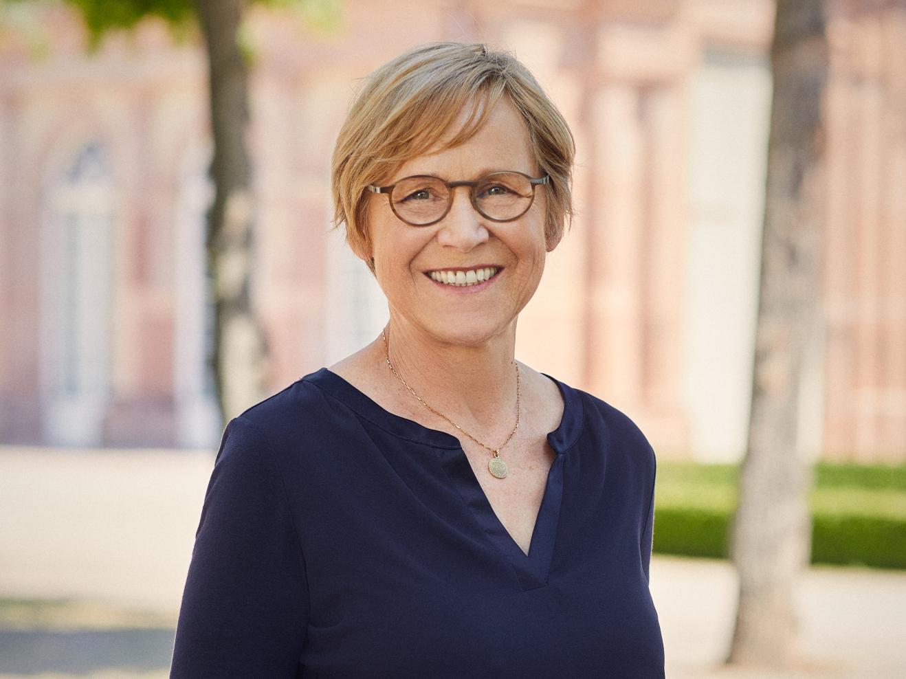 Portraitfoto von Heike Hoffmann. Sie hat kurze blonde Haare, trägt eine Brille und lächelt in die Kamera. Ihre Hände hat sie in den Hosentaschen. Sie trägt eine blaue Bluse, eine blaue Hose und eine goldene Kette.