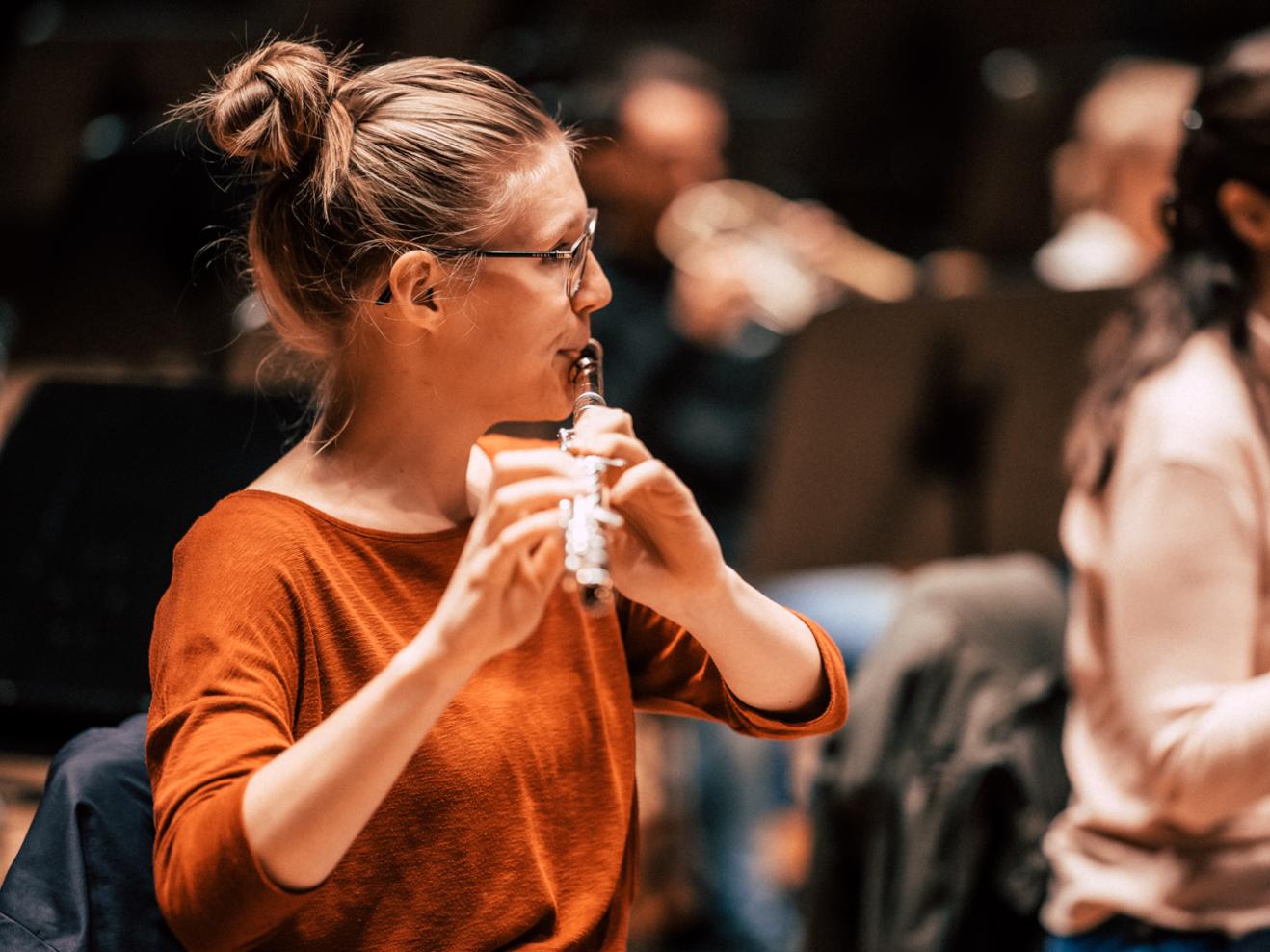 Sina Merkel spielt Querflöte in einer Orchesterprobe. Sie trägt ein organgenes Shirt, eine Brille und hat ihre dunkelblonden Haare zu einem Dutt gebunden. Man sieht sie hier im Profil.