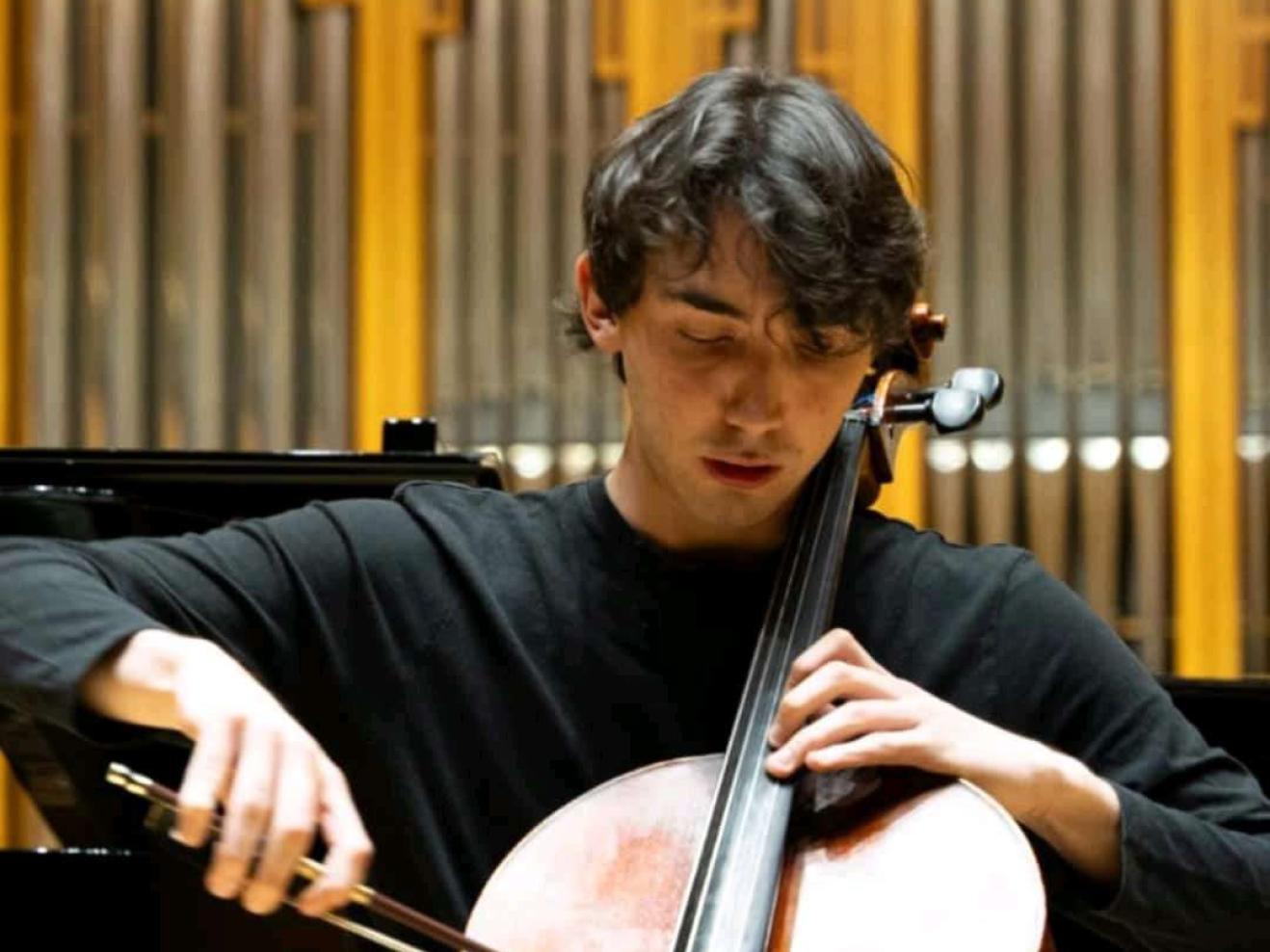 Unai Sánchez spielt Cello. Er hat kurze, dunkle Locken und blickt hinab auf die Saiten. Er trägt eine graue Hose und einen schwarzen Pullover. Im Hintergrund zu sehen sind ein schwarzer Flügel und die Pfeifen einer Orgel.