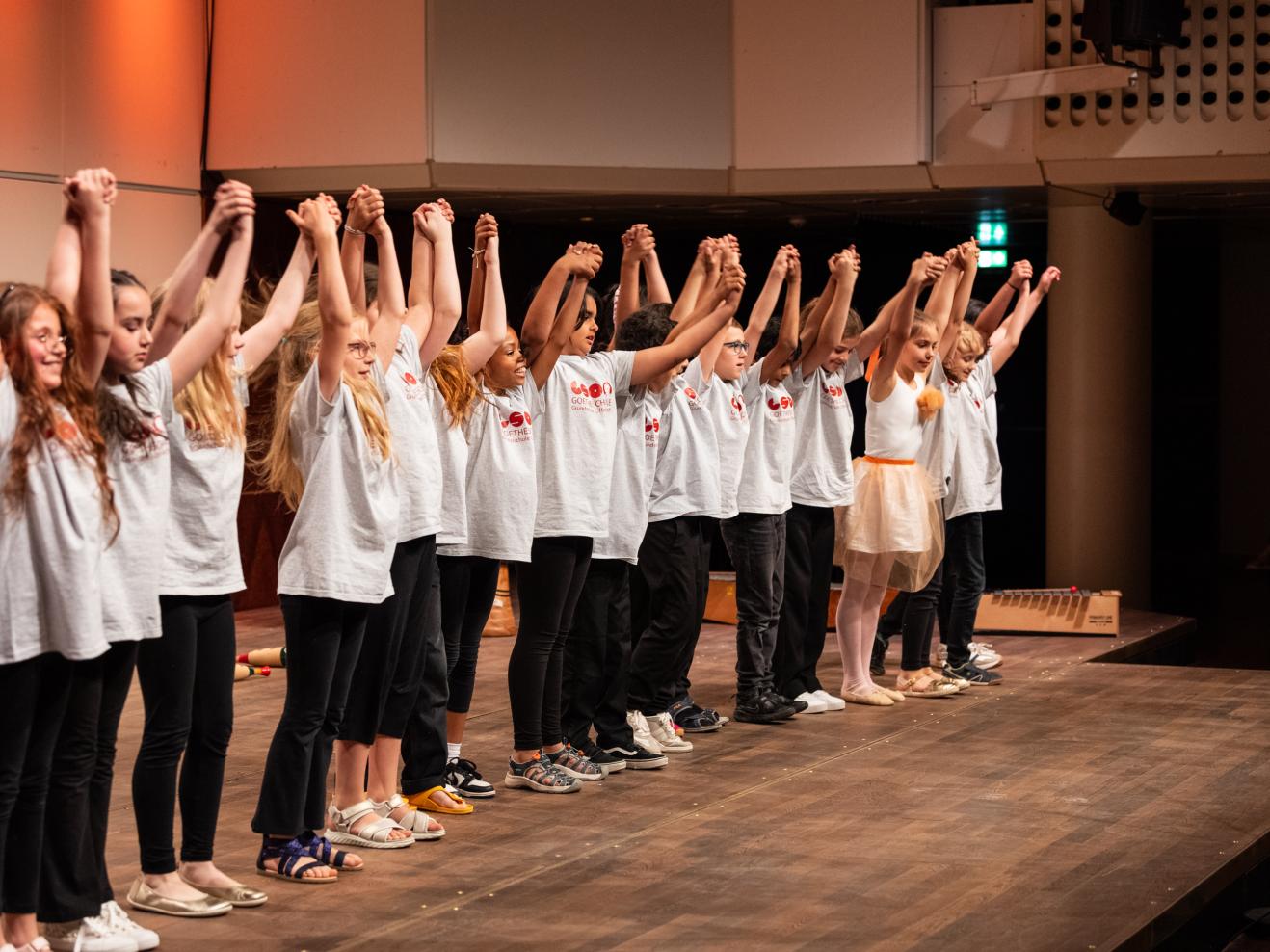 Eine Reihe von Kindern in weißen T-Shirts steht auf der Bühne und verbeugt sich. Alle fassen sich an den Händen und haben die Arme nach oben gestreckt.