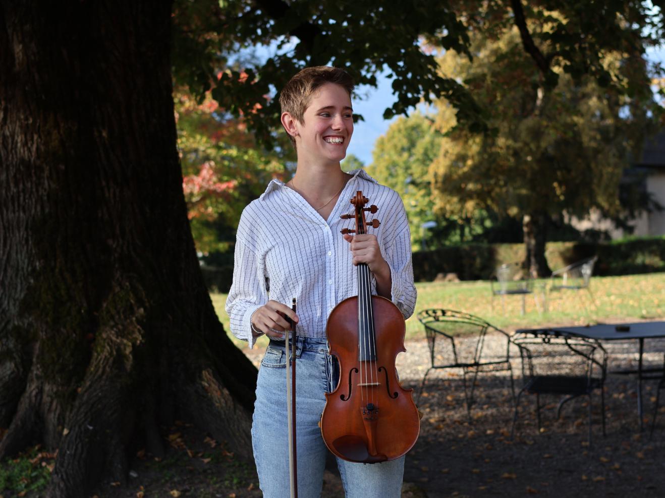 Tamara Durand steht lachend vor einem Baum und hält ihre Viola in der Hand.